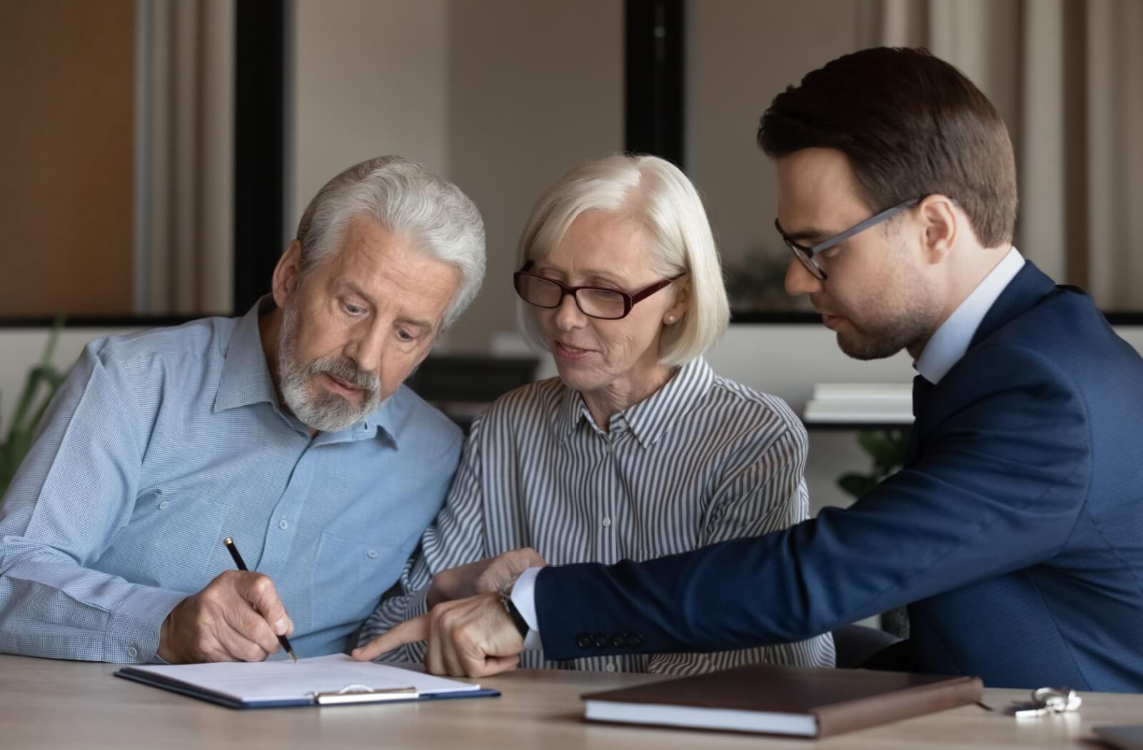 A lawyer sitting at a table with an older couple while the older man signs a document the lawyer is pointing at.