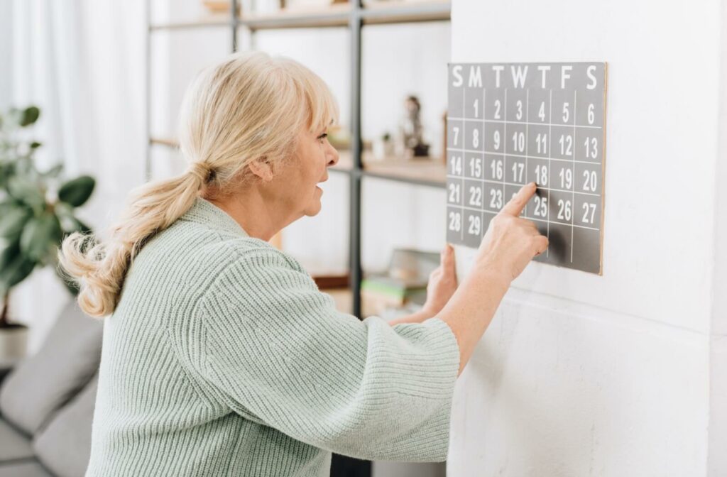 An older woman looking at a calendar on the wall and pointing to a date on the calendar.