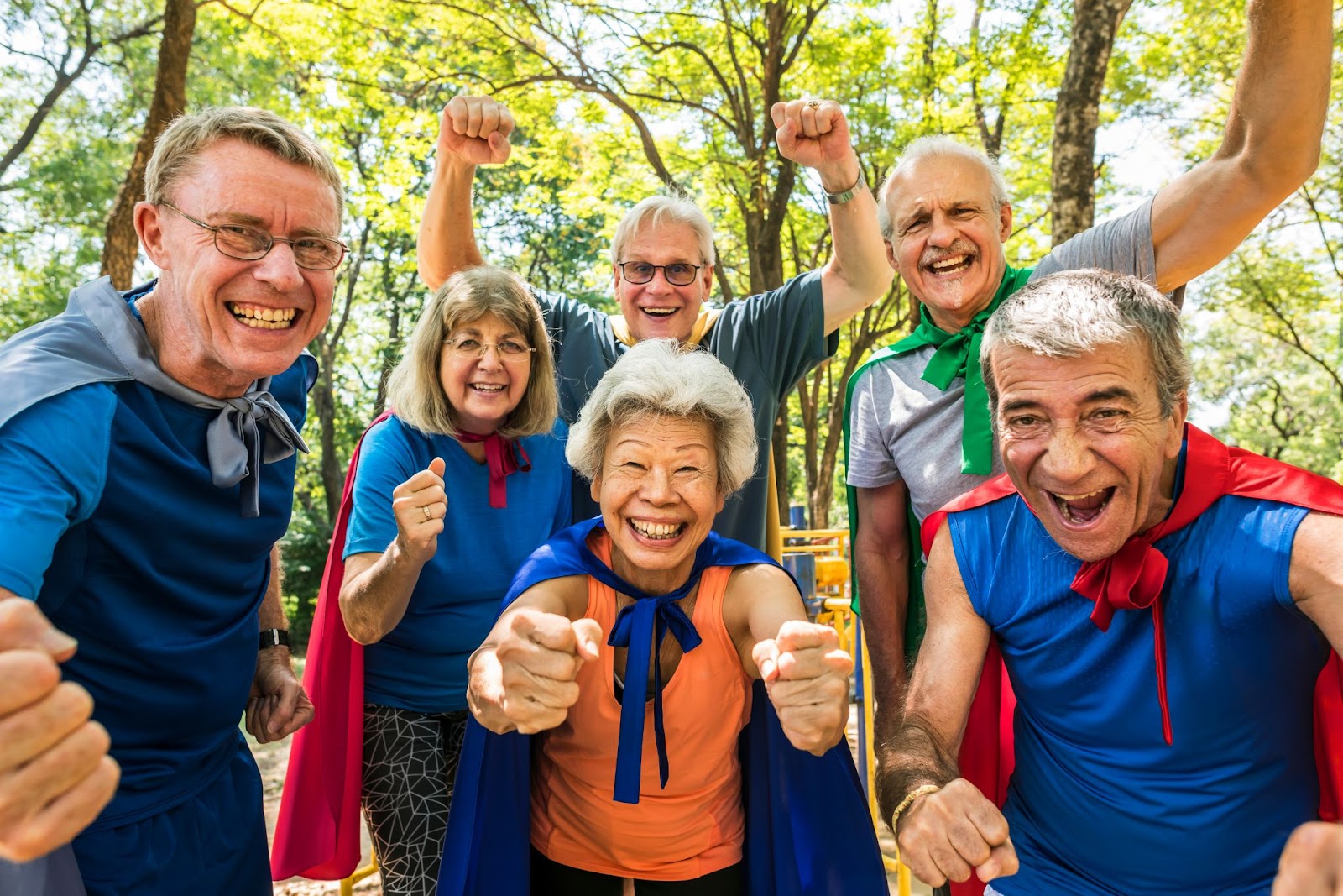 A group of several seniors wearing superhero capes in the park in the daytime, supporting each other as a community