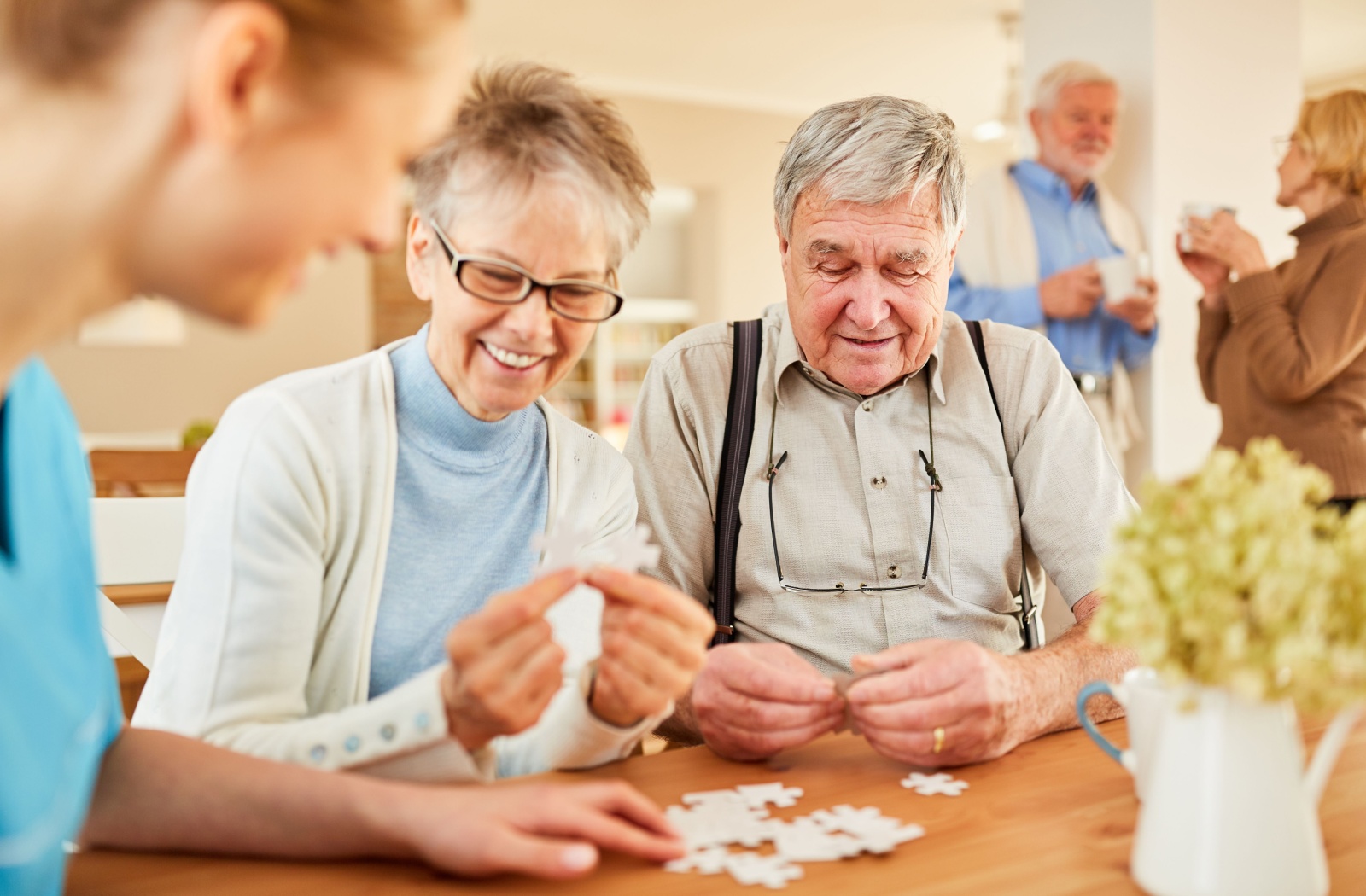 Two older adults working on a puzzle with the help of a caregiver, while others chat in the background.