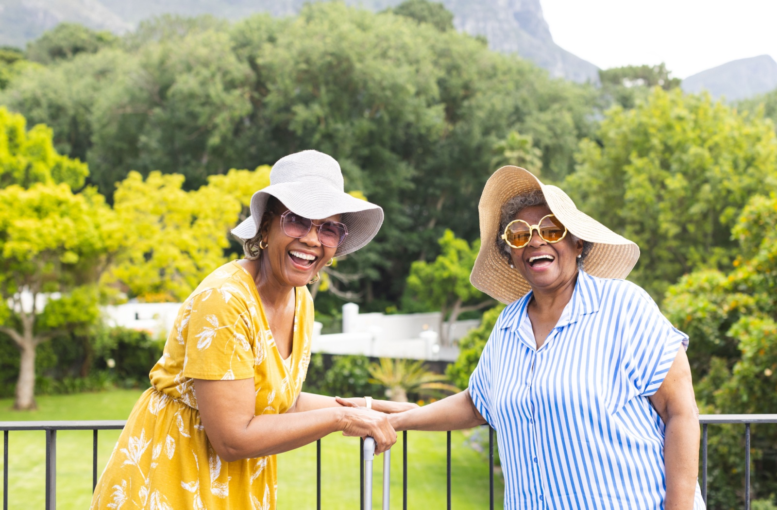 Two senior women sharing a laugh on a sunny balcony, both wearing wide-brimmed hats for sun protection.