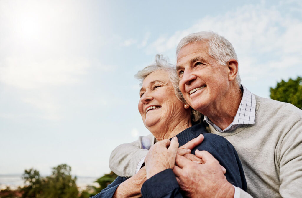 An older adult hugging their spouse from behind while on a walk together outdoors in the summer.