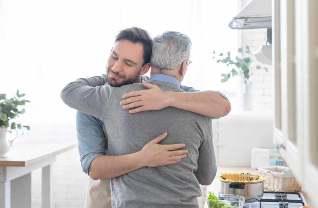 An adult child hugging their older parent in the kitchen while making dinner and talking about memory loss.