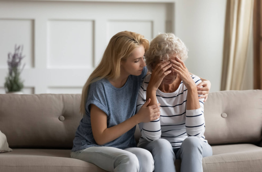 An adult child hugs their older parent to reassure them while sitting together on the couch and talking about a potential move to memory care