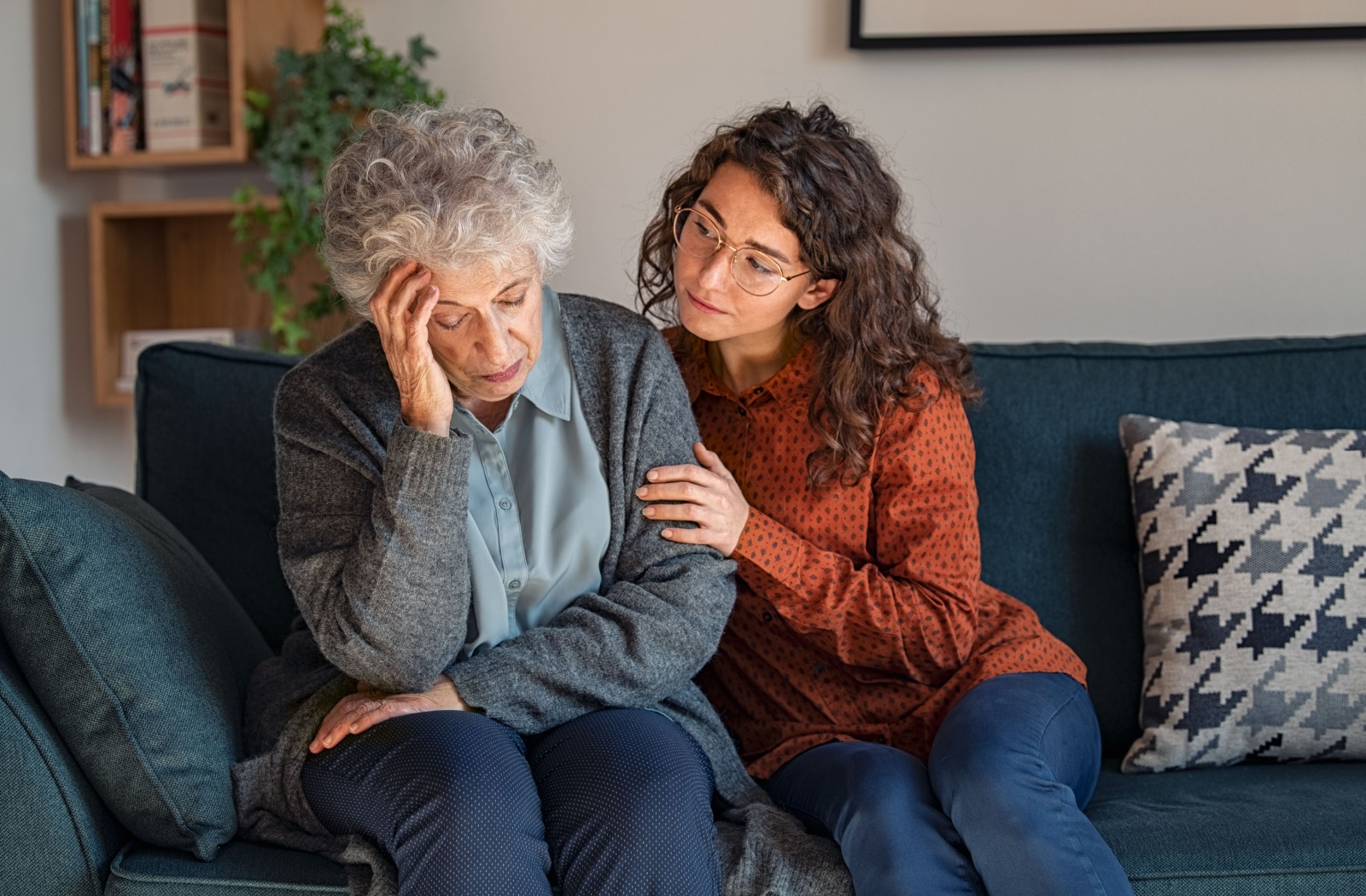 An adult child comforts their distressed older parent on a navy blue couch during a conversation about memory care in their living room